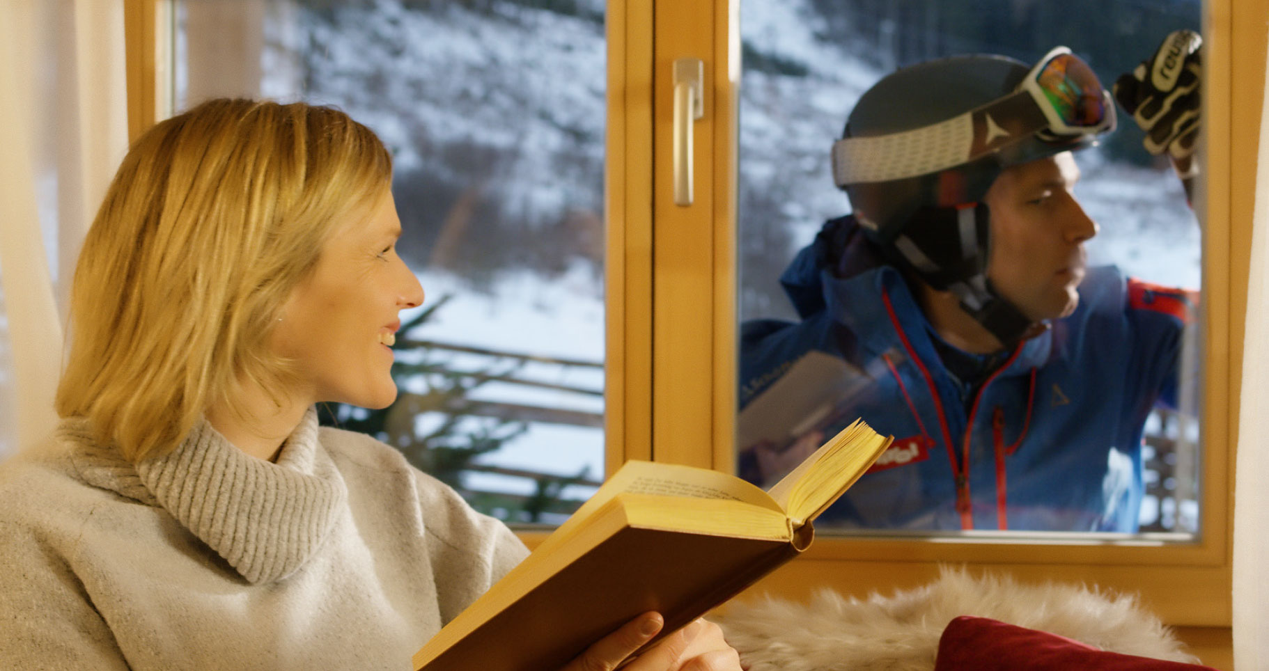 Frau mit Buch vor Fenster, Mann mit Skiausrüstung dahinter