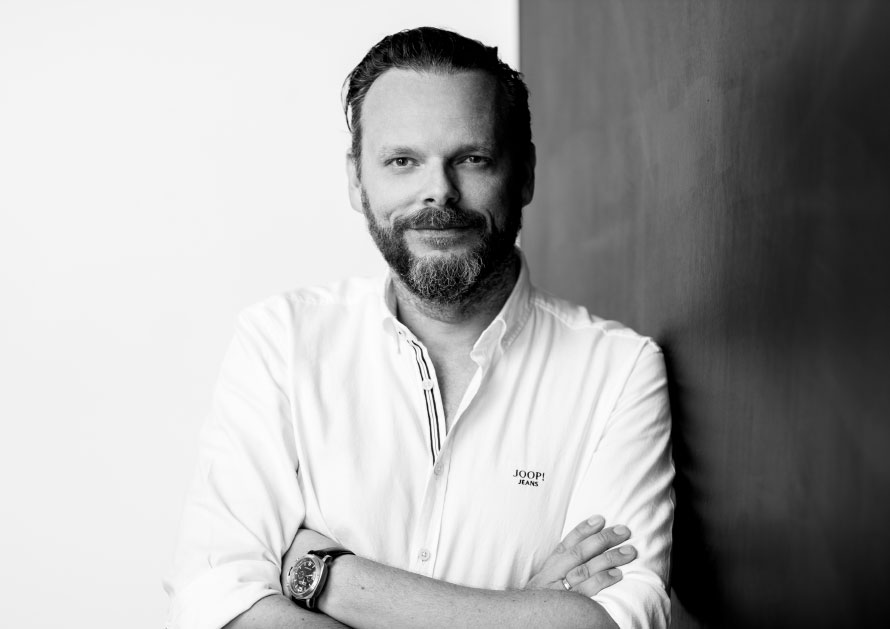 Jürgen Mellak, Mann mit Drei-Tages-Bart und weißem Hemd sowie Armbanduhr, lehnt an eine Wand